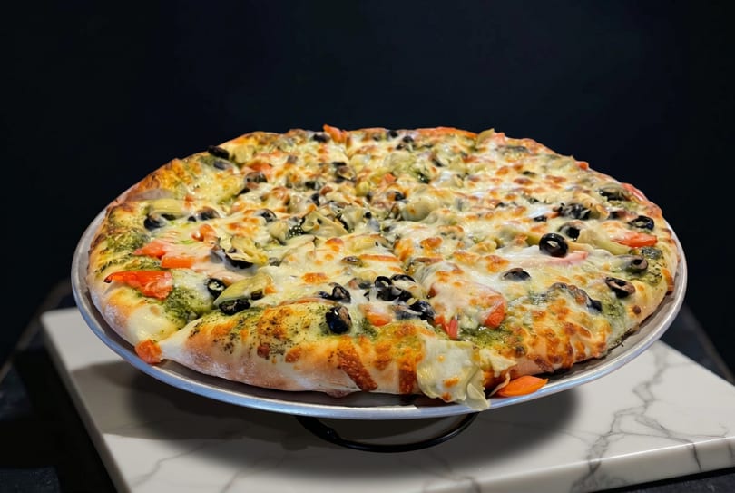 A whole pizza with pesto, cheese, tomatoes, olives, and artichoke hearts on a metal pan.